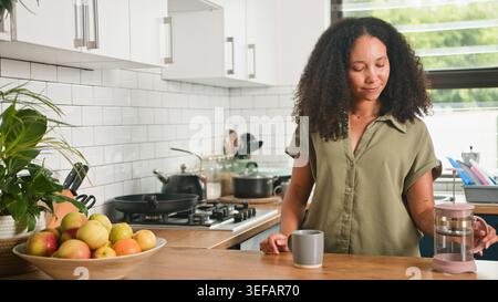 Junge Frau, die Kaffee in der modernen Küche mit Obstschale und entspannter Morgenstimmung macht Stockfoto