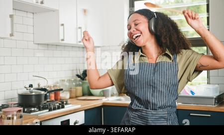 Happy Young Woman tanzt in der modernen Küche beim Kochen, trägt Kopfhörer und gestreifte Schürze Stockfoto