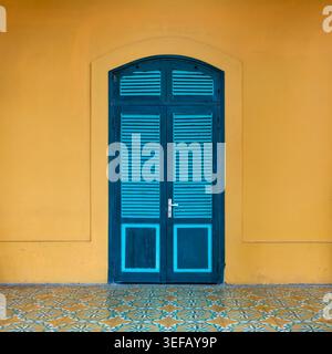 Leuchtend blaue Lamellentür mit gewölbter Oberseite und silbernem Griff, vor einer hellgelben Wand, über verzierten gemusterten Bodenfliesen, traditionelle Architektur, Hanoi, Vietnam Stockfoto