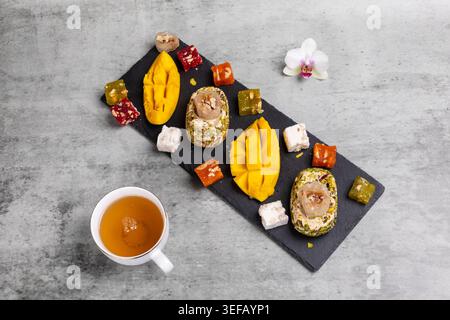 Tasse Tee. Halva mit Pistazien, einer geschnittenen frischen Mangobrucht und Fruchtsüßen mit Nüssen auf dem Schneidebrett aus Stein. Draufsicht. Stockfoto