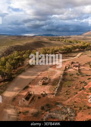 Aus der Vogelperspektive auf die historischen Ruinen der Kanyaka Station und eine gewundene, unbefestigte Straße, umgeben von trockener Landschaft und Bäumen unter einem bewölkten Himmel in den Flinders Ranges Stockfoto