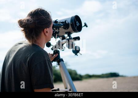 Nahaufnahme von hinter dem Mann, der durch ein Teleskop am Strand beobachtet. Shot konzentriert sich auf seine Haltung und das Teleskop und betont die Konzentration und den technischen Aspekt der Sonnenbeobachtung. Stockfoto