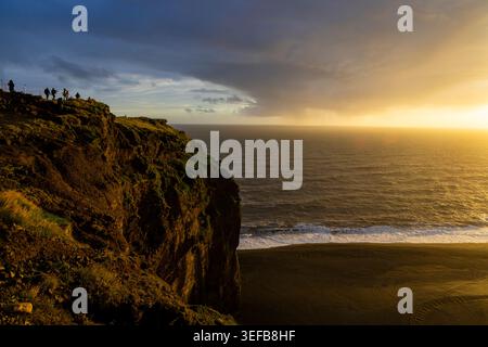 Goldenes Sonnenlicht leuchtet auf Klippen und Küsten am Dyrholaey Beach in Island, während Wellen gegen schwarzen Sand krachen Stockfoto