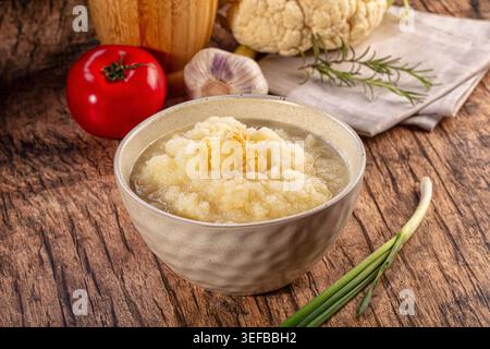 Eine warme, rustikale Szene mit einer Keramikschale mit cremigem Blumenkohlpüree. Frische Zutaten wie Tomaten, Knoblauch, Rosmarin umgeben das Gericht auf einem Holz Stockfoto