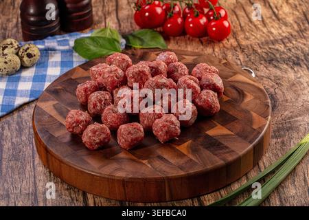 Eine Nahaufnahme roher Fleischbällchen auf einem rustikalen Holzschneidebrett, umgeben von frischen Zutaten wie Kirschtomaten, Basilikum, Wachteleiern usw. Stockfoto