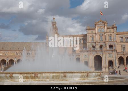 Zentraler Brunnen mit Wasserstrahl vor dem reich verzierten Hauptpavillon und der spanischen Flagge an der Plaza de España, Sevilla, Andalusien, Spanien. Stockfoto