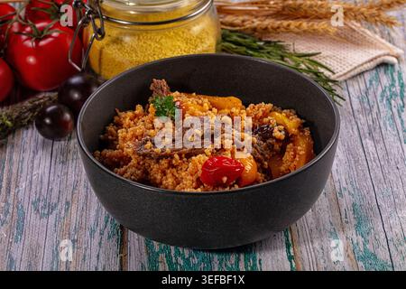 Herzhaftes Couscous-Gericht mit zartem Rindfleisch und Gemüse in dunkler Schüssel. Rustikale Speisen mit frischen Tomaten, Kräutern und rohem Getreide auf dickem Holz Stockfoto