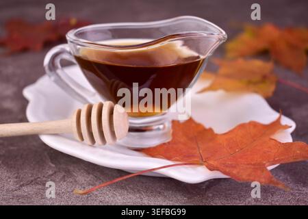 Süßer Ahornsirup in einem Glas Soße Boot und Ahornblätter auf grauem Hintergrund. Stockfoto