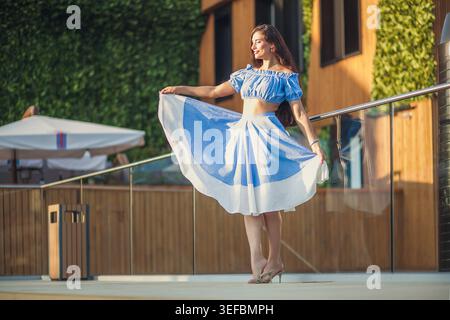 Junge glückliche Frau mit langen Haaren in stilvollem blau-weißem Sommeroutfit. Sie lächelt und wirbelt ihren fließenden Rock auf der modernen Terrasse Stockfoto