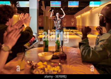 Junge Erwachsene Frau, die Bowling feiert, gewinnt, während Freunde in der Gasse vom Tisch klatschen und Teamarbeit und Freizeit zeigen. Ideal für gesellschaftliche Veranstaltungen, Gruppenausflüge, Freizeitmarketing Stockfoto