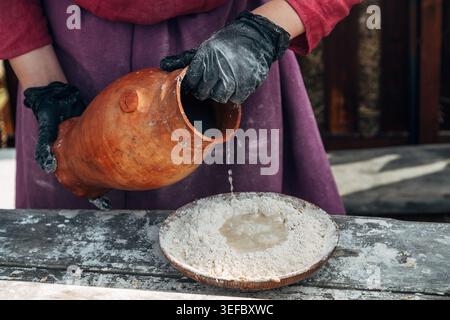Frauenhände gießen Wasser aus einem Tonkrug, um Teig nach einem alten russischen Rezept zu kneten. Stockfoto