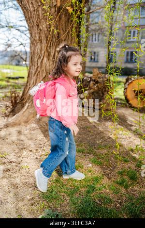 Weißes Mädchen mit rosafarbenem Rucksack, das in der Nähe von Baumzweigen im Wohnpark spaziert. Kindheit und Outdoor-Lifestyle. Stockfoto
