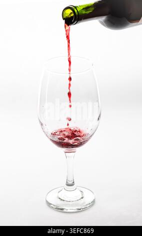Rotwein wird aus einer Flasche in ein transparentes Weinglas vor weißem Hintergrund gegossen. Elegante Getränkefotografie, die flüssige Bewegungen einfängt. Stockfoto