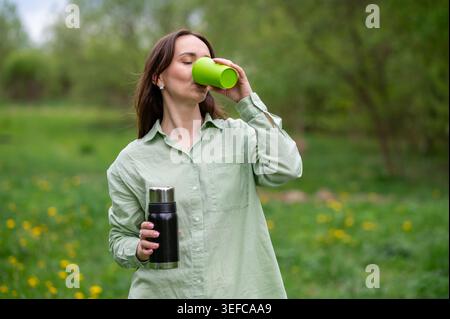 Eine junge kaukasische Frau trinkt ein warmes Getränk aus einer grünen Tasse, während sie eine schwarze Thermoskanne hält, während sie in einem wunderschönen grünen Park mit b spaziert Stockfoto