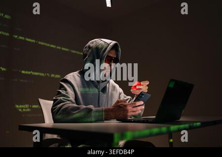 Gefährlicher Hacker mit Kapuze, männlich mit Sonnenbrille, mit Smartphone und roter Kreditkarte, umgeben von grünen Codes, die auf Cyberkriminalität und Betrug hinweisen Stockfoto