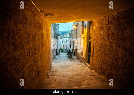 Beschreibung: Ein Blick durch eine gewölbte Steinpassage im historischen Zentrum von Ragusa Ibla, Sizilien. Die Perspektive blickt auf eine steile Kalksteintreppe Stockfoto