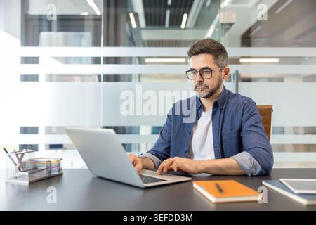 Ein fokussierter Mann mit Brille und Denim-Hemd, der an einem modernen Schreibtisch sitzt und fleißig auf seinem Laptop tippt, was Produktivität und professionelle Arbeitsumgebung verkörpert Stockfoto