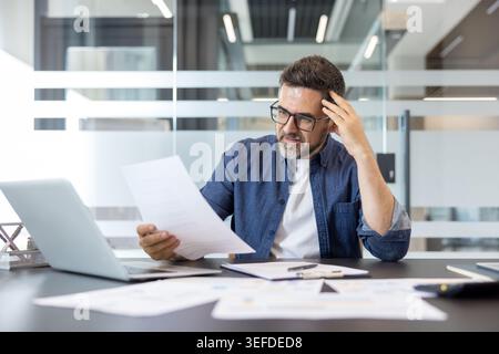 Reifer Geschäftsmann in einem modernen Büro, der sich mit Finanzberichten und Papieren beschäftigt, sichtbar gestresst und besorgt bei der Datenanalyse, Termindruck und schwierigen Entscheidungen ausgesetzt ist Stockfoto