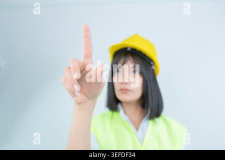 Junge Bauarbeiterin mit gelbem Schutzhelm und reflektierender Sicherheitsweste, die mit Fingergesten nach oben zeigt. Studio isoliert mit Licht aufgenommen Stockfoto