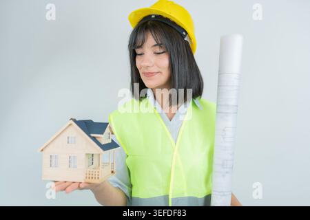 Architektin oder Ingenieurin mit Miniatur-Hausmodell und Blaudruckrolle, während sie einen Schutzhelm und eine reflektierende Weste trägt. Studio-Fotografie repräsentieren Stockfoto