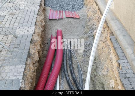Installation von Erdkabeln und -Rohren in städtischen Versorgungsgräben. Schwarze Elektrokabel und rote Wellrohre sind in einem Straßengraben sichtbar Stockfoto