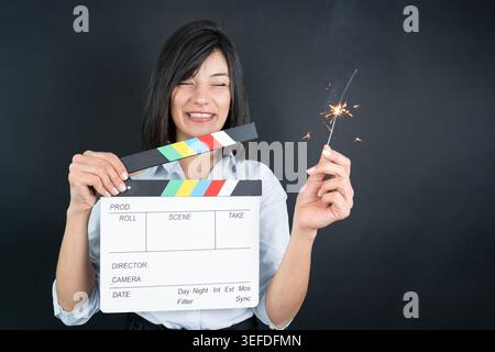 Lächelnde Frau mit Filmklappertafel und funkelndem Feuerwerk in Studiobeleuchtung. Konzept von Filmgestaltung, Unterhaltung, Feier und kreativer Medizin Stockfoto