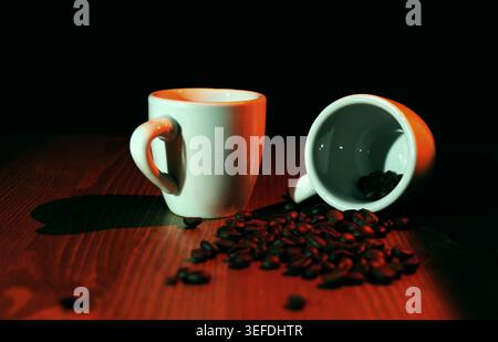 Zwei weiße Espresso-Tassen aus Keramik auf einem dunklen Holztisch, warmes, stimmungsvolles Licht, das geröstete Kaffeebohnen aus einer umgedrehten Tasse hervorhebt, einladend Stockfoto