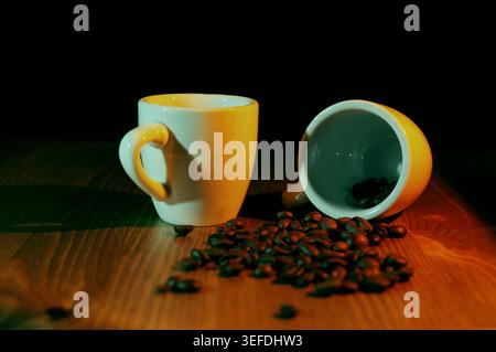 Kaffeebohnen, die aus einer umgedrehten Espresso-Tasse neben einer stehenden Tasse austreten, schaffen ein einladendes Stillleben auf einer Holzoberfläche mit warmem und kühlem Li Stockfoto