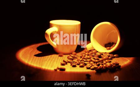 Kaffeebohnen auf einer Holzoberfläche neben zwei kleinen Espresso-Tassen verstreut, von denen eine steht und eine umgekippt ist, was eine warme, einladende Szene in einem Gentlemen schafft Stockfoto
