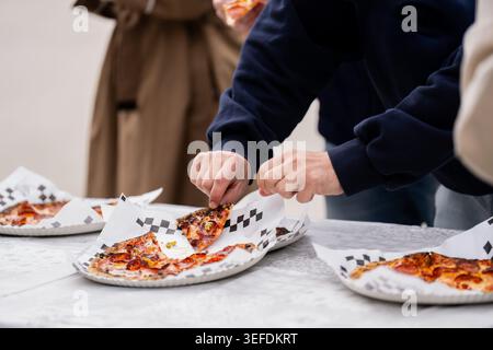 Hungrige Freunde, die bei einem zwanglosen Treffen auf einem weißen Tisch nach frischen Pizzabschnitten mit roten Belägen greifen und dabei den Lebensstil junger Menschen und schnellen Foo hervorheben Stockfoto