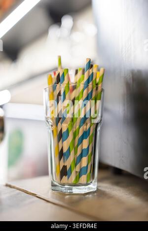Bunte gestreifte Papierhalme in einem klaren Glas. Glas sitzt auf einer Holzoberfläche. Hintergrund zeigt verschwommene Küchengeräte. Sanfte Lichter Stockfoto