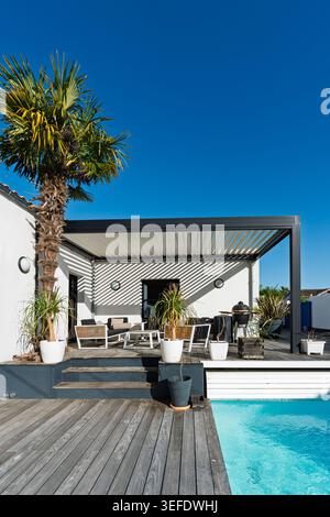 Flacher Blick auf eine Palme, die sich gegen eine bioklimatische Pergola aus schwarzem Aluminium erhebt, mit einer Holzterrasse, übertöpften Pferdeschwanz-Palmen und einem Lounge-Bereich im Freien. Stockfoto