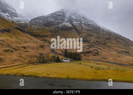 Aus der Vogelperspektive auf das weiße Häuschen und die schneebedeckten Berge in der zerklüfteten Landschaft von Glencoe, Schottland, Großbritannien. Stockfoto