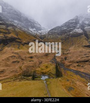 Blick aus der Vogelperspektive auf ein kleines weißes Cottage am Fuße dramatischer schneebedeckter Berge und zerklüfteter Tallandschaft in Glencoe, Schottland, United King Stockfoto