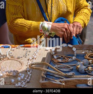 Frau in einem gelben Pullover inspiziert handgemachten Schmuck auf einem Holztisch auf einem Flohmarkt und stellt eine Vielzahl von Ketten und Armbändern vor Stockfoto