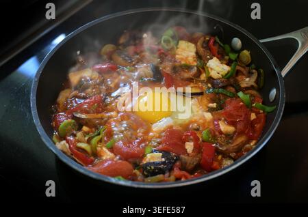 Gemüse-Shakshuka in einer dampfenden schwarzen Pfanne, hausgemachtes Rezept mit Tomaten, Paprika, Auberginen, Zwiebeln, Lauch, Feta und ein pochiertes Ei, Levantinkochen Stockfoto