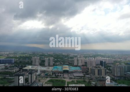 Unter einem dynamischen Himmel entfaltet sich eine weitläufige moderne Stadtlandschaft, in der strahlende Lichtstrahlen durch dichte Wolken brechen und ein hohes Gebäude erleuchten Stockfoto