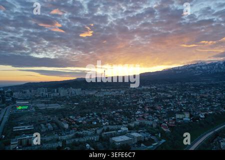 Eine Luftperspektive fängt eine weitläufige urbane Landschaft ein, die in warmes goldenes Licht getaucht ist, während die Sonne hinter entfernten, schneebedeckten Bergen untergeht Stockfoto