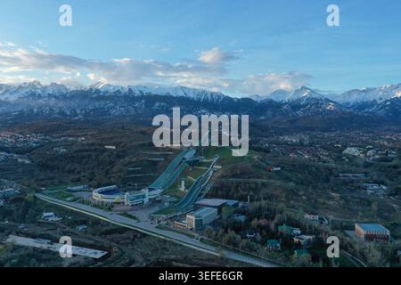 Eine moderne Skisprungschanze, eingebettet in sanfte Hügel mit schneebedeckten Bergen, die majestätisch hinter einem klaren blauen Himmel ragen. Stockfoto