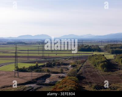Luftaufnahme von Hochspannungsleitungen und Strommasten durch landwirtschaftliche Felder und ländliche Landschaft mit Bergen in der Ferne Sloweniens Stockfoto