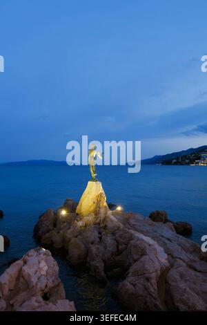 Das beleuchtete Mädchen mit der Möwenstatue an der felsigen Küste von Opatija, Kroatien, während der blauen Stunde Stockfoto