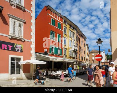 Menschenmassen laufen durch eine sonnige Straße mit farbenfrohen historischen Gebäuden und Restaurants im Freien in Rovinj, Kroatien, Europa Stockfoto