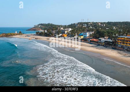 Aus der Vogelperspektive auf die Sandküste von Kovalam Beach mit türkisfarbenen Wellen, die an der Küste zusammenbrechen und Palmen, die die Gebäude am Wasser säumen, unter A Stockfoto