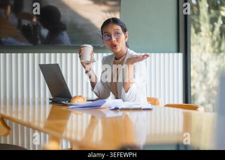 Lächelnde Geschäftsfrau in Brille sitzt an einem hölzernen Besprechungstisch mit Laptop, Stift und gedruckten Verkaufsberichten. Sie zeigt die Unternehmensplanung, die Finanzplanung Stockfoto