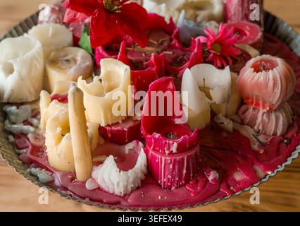 Mehrere geschmolzene rot-weiße Wachskerzen mit dekorativen Blumen sitzen in einem Metalltablett auf einem rustikalen Holztisch in einem Wohnraum. Stockfoto