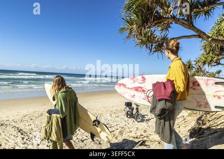 Zwei Frauen mit Surfbrettern gehen an einem sonnigen Strand an der Goldküste in Richtung des glitzernden pazifiks und genießen Urlaubsstimmung, warmes Wetter und unbeschwerte Surf Re Stockfoto