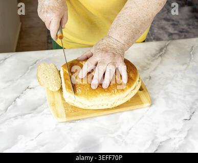 Frau schneidet frisch gebackenes hausgemachtes Brotbrot mit Messer auf hölzernem Schneidebrett auf Marmorküche, Studio-Aufnahme Stockfoto