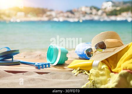 Sonnenhut, Sonnenbrille, Sonnencreme, Handtuch und Strandspielzeug sind am Sandstrand mit türkisfarbenem Meer und sonniger Kulisse an der Küste angeordnet. Stockfoto
