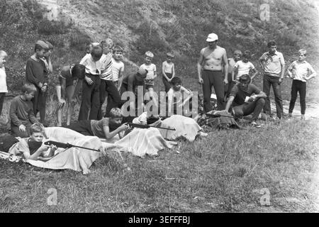 Sloviansk, 1983. Slavkurort, in der Nähe von Wishnegorsk Pionierlager. Ein schwarzweißes Archivfoto, das eine Gruppe sowjetischer Kinder zeigt, die in einer grasbewachsenen Schlucht mit Luftgewehren Schusskunst üben. Die Jungen und Mädchen liegen auf Decken und zielen vorsichtig auf Ziele, während Erwachsene Instruktoren die Sommeraktivitäten überwachen. Andere Kinder stehen im Hintergrund und beobachten den Prozess. Dieses authentische Vintage-Bild zeigt die Sportausbildung im Freien und die alltägliche Freizeit der Jugendlichen während der Zeit des friedlichen Donbass. Stockfoto