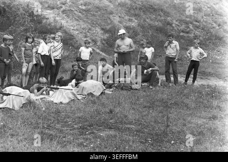 Sloviansk, 1983. Slavkurort, in der Nähe von Wishnegorsk Pionierlager. Ein schwarzweißes Archivfoto, das eine Gruppe sowjetischer Kinder zeigt, die in einer grasbewachsenen Schlucht mit Luftgewehren Schusskunst üben. Die Jungen und Mädchen liegen auf Decken und zielen vorsichtig auf Ziele, während Erwachsene Instruktoren die Sommeraktivitäten überwachen. Andere Kinder stehen im Hintergrund und beobachten den Prozess. Dieses authentische Vintage-Bild zeigt die Sportausbildung im Freien und die alltägliche Freizeit der Jugendlichen während der Zeit des friedlichen Donbass. Stockfoto